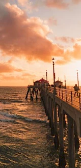 A sunset view of a pier extending into the ocean, with a few people walking along it and buildings at the end, under a sky filled with clouds.